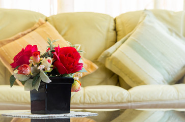 Beautiful artificial flowers in vase on mirror table with sofa and backrest pillow background in living room. 