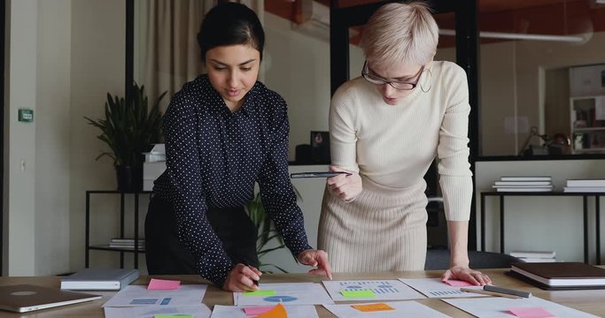 Diverse Young Business Women Brainstorm On Project Plan Paperwork Applying Sticky Post It Notes On Table. Two Indian And Caucasian Coworkers Discuss Business Strategy Put Stickers At Office Meeting.