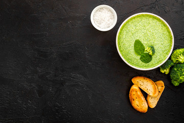 Broccoli cream soup, bread, spoon on black kitchen desk top-down copy space