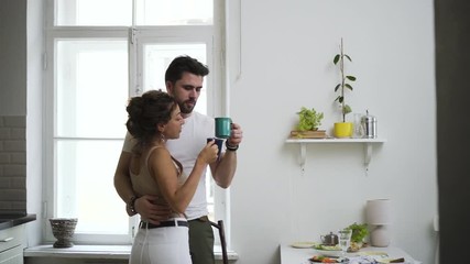 Young woman and bearded man are drinking coffee and kissing while standing in white interior spbd. Handsome guy and pretty female enjoy drink, kiss and hug, having good morning in modern apartment - Powered by Adobe
