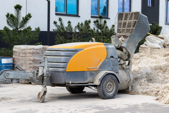 Modern Concrete Screed Pump At A Construction Site Next To A Pile Of Sand, Bags Of Cement, A Building And Pine Trees