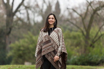 Beautiful Indian woman dressed in poncho smiling in park. Looking at camera.