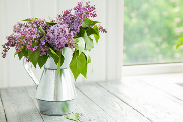 metal watering can with a bouquet of lilac on a wooden background near the window