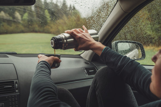 Female pouring the hot tea in tourist thermos mug. She sitting on co-driver seat inside modern car, enjoying the moody rainy day weather looking through windscreen drops. Auto journeys concept image.