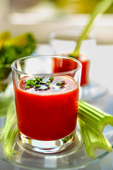 Two glasses of tomato juice with parsley and celery decorations, stand near the window, morning sunlight shines, shallow depth of field, selective focus. Natural drinks concept.
