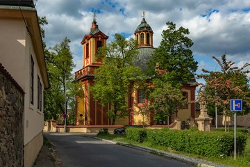 Fototapeta na wymiar Kunratice, Prague / Czech Republic - May 6 2020: The baroque church of St James the Great with red and yellow facade surrounded with green trees. Sunny day with blue sky and white clouds.