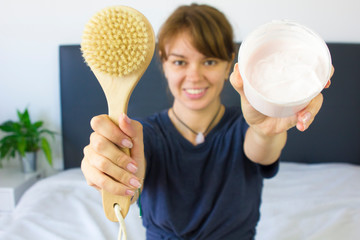 Beautiful fit young ginger woman at home in bed in pajama. She show items for beauty procedures- dry massage eco comb. and moisturizing cream. Lifestyle home spa healthcare concept