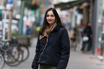 Beautiful young middle eastern woman listening to earphones smiling at camera on sidewalk.