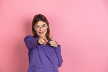 Pointing, choosing. Caucasian young woman's portrait isolated on pink studio background. Beautiful female model in purple jacket. Concept of human emotions, facial expression, sales, ad. Copyspace.