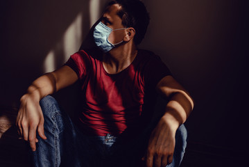 Fototapeta na wymiar Sad thoughtful adult man sideview profile portrait in medical mask sit leaning wall with light and shadow pattern. Stay isolation at home. Quarantine concept. Pandemic Coronavirus. Social Distancing