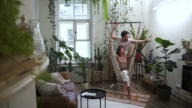 middle age romantic couple in beautiful hipster home apartment interior with plants taking selfies on smart phone Spbd. woman sitting in hanging swing. relationship, married, technology concept