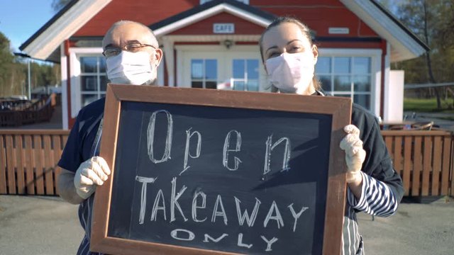 A middle-aged couple wearing medical masks standing in front of their restaurant and holding a "takeaway only" sign. Small business during coronavirus quarantine