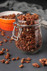 Granola crispy muesli with natural honey, chocolate, nuts in a glass jar and bowl against a dark background, vertical format