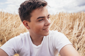 Close-up portrait of cheerful handsome teenage boy, sitting on wheat field, under a sky with...