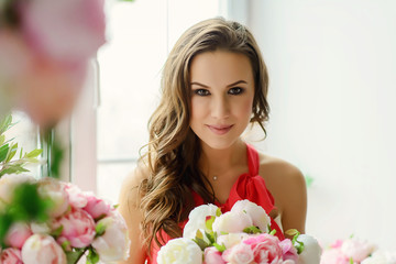 Beautiful young brown-eyed woman with a bouquet of peonies flowers in her hands near the window.