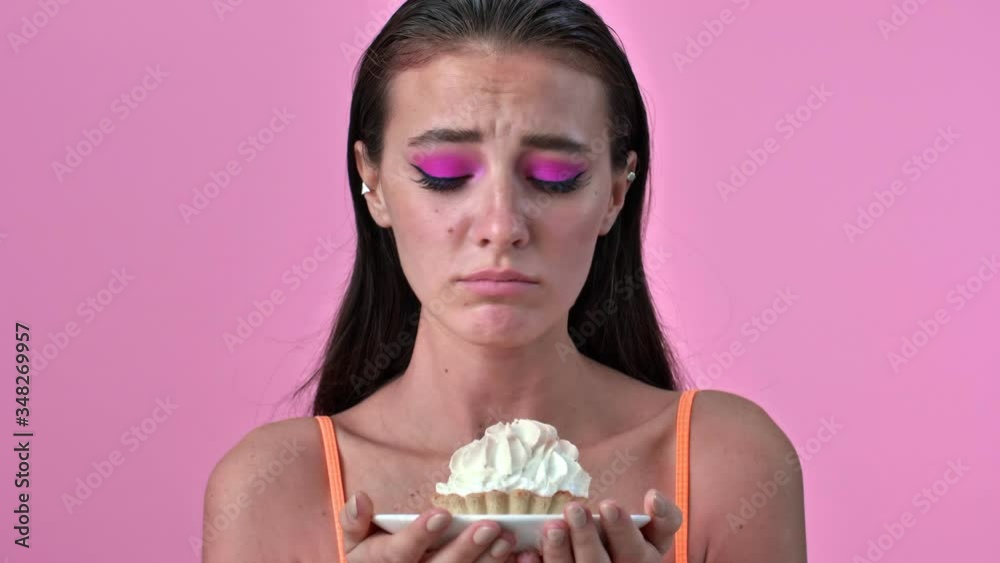 Wall mural Young hungry woman isolated over pink wall background holding cupcake