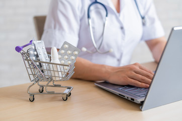 Female doctor working on a laptop. Mini shopping trolley full of pills. Online pharmacy concept. A...