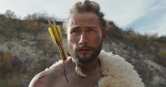 Bearded man in animal hide and with arrows looking around while standing near cliff on autumn day in wilderness.