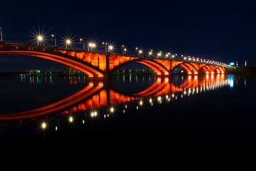 Communal bridge over the Yenisei River in Krasnoyarsk, Russia. Reflection red illuminations in the dark water. Bright lights of night city.