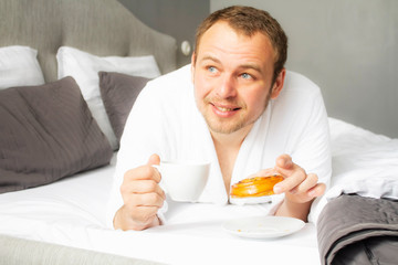 Handsome young man having breakfast while lying in bed. The guy on the white bed with food. Breakfast in bed.