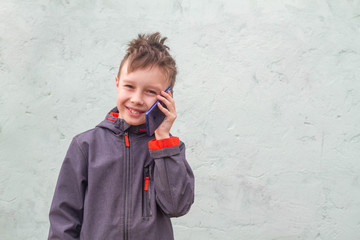 A teenage European boy stands against the wall of the house and learns about the news on a blue smartphone. The child is happy and smiles.