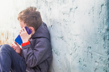 A teenage European boy sits against the wall of the house and learns about the news on a blue smartphone. Difficult conversation.