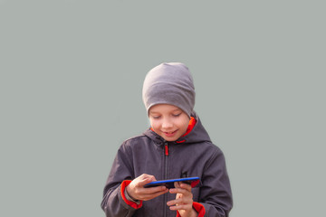 A European teen boy is playing a game on his smartphone. A child in a gray hat and blue jacket on a gray background.