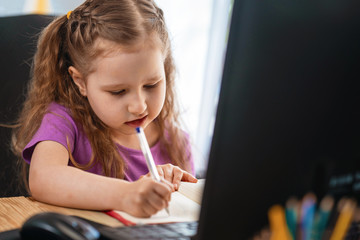 A little cute girl using computer for remote e-learning, , writes in a notebook