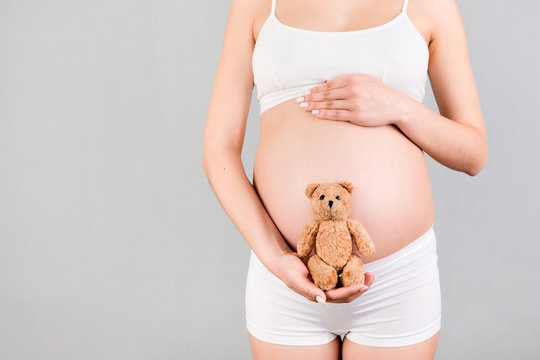 Cropped image of happy pregnant woman in white underwear holding teddy bear against her belly at gray background. Child expecting. Copy space