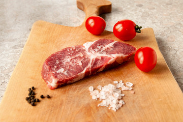 fresh raw steak lies on a cutting board, next to salt, pepper, tomatoes