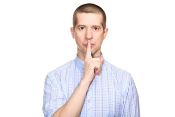 Serious man in a classic blue shirt; shows a gesture of silence, the preservation of silence and mystery; put his index finger to his lips; look into the camera. Isolated
