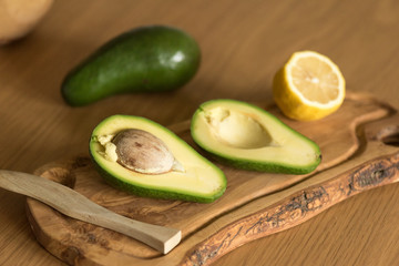 Two halves of avocado and half a lemon on an olive board, another whole avocado and a bowl on the...