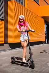 Fototapeta na wymiar Beautiful, slender, young girl riding an electric scooter, near a shopping center, having fun, laughing, listening to music, denim shorts, hat, fashionable top