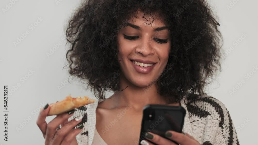 Wall mural Young happy cheery positive african curly woman indoors at home using mobile phone eat pizza