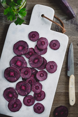 Overhead view on slices of raw purple carrot on a white marble board over kitchen table.