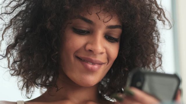 Young positive african curly woman indoors at home using mobile phone