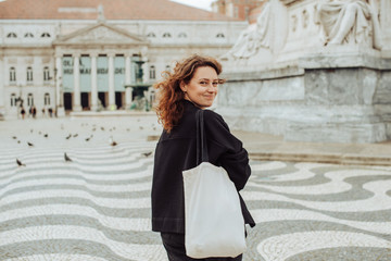 Half length portrait of caucasian millennial woman looking back over her shoulder and smiling at camera. Posing on Rossio Square in Lisbon, Portugal. Copy space
