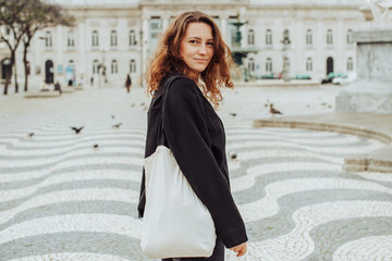 Half length portrait of caucasian millennial woman looking back over her shoulder and smiling at camera. Posing on Rossio Square in Lisbon, Portugal with traditional Portuguese pavement