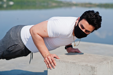 Portrait sports arabian man in black medical face mask doing morning workout exercises against lake during coronavirus quarantine.