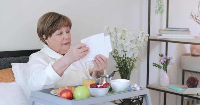 Mature good looking lady looking photos during breakfast in bed. Elderly woman in home wear smiling while sitting with trey full of food and going through family photos.