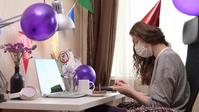 Woman celebrating her birthday through video call virtual party while wearing mask. Lits and blows out candle. Authentic decorated home workplace. Handheld shot with gimbal. Coronavirus outbreak 2020.