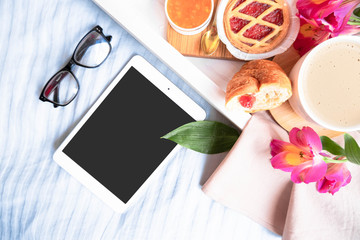 Home learning. Online education. Top view. Breakfast with croissant, cup of coffee, cappuccino, flowers, jam. Woman`s learning. Tablet and glasses on bed. Freelance. Tablet mockup. Work place