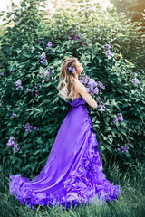 Obraz na płótnie Canvas A beautiful young woman enjoys the smell in a blooming spring Lilac garden. Flowers in her hair. She's wearing a long, chic purple dress with a train. Artistic image of high fashion