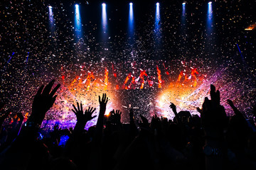 Fototapeta Fans hands raised up during the show. Bright lights and shiny confetti at a pop concert. happy youth dancing at a festival in a crowd.  view of the stage. obraz