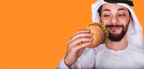 Close-up of  Arab man and a burger. Arabic guy closes his eyes and smells the scent of a delicious fresh burger on orange background.Delicious fast food concept. Copyspace