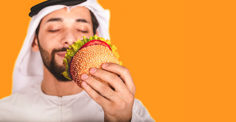 Close-up of  Arab man and a burger. Arabic guy closes his eyes and smells the scent of a delicious fresh burger on orange background.Delicious fast food concept. Copyspace