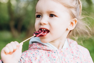 little 3-year-old girl licks a colored swirling candy on a walk in the park on a rainy summer day