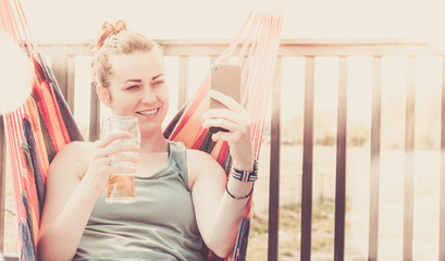 Smiling woman relaxing on a hammock - Talking on a video communicator and drinking a beer - Concept...