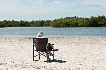Fototapeta na wymiar Back view, medium distance of a senior woman relaxing in a beach chair on a sandy beach off the coast of an island on gulf of Mexico on a sunny, spring day