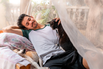 hombre joven recostado en un sillon en el jardin detrás de una mosquitera, acaricia un gato negro sentado en su costado